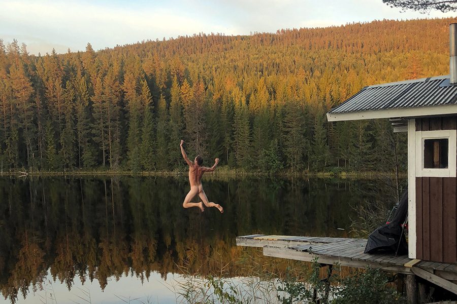 En man som hoppar naken ut i vattnet från bryggan vid en bastu, alldeles vid vattenbrynet.