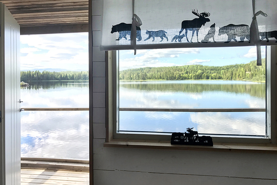 Inne i en stuga, ett fönster ut mot sjön alldeles utanför. Vattenytan är alldeles blank, en solig sommardag.