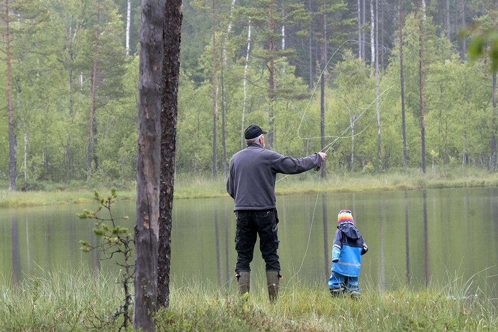 En man och en liten pojke i klarblå regnkläder fiskar vid en liten tjärn i skogen.